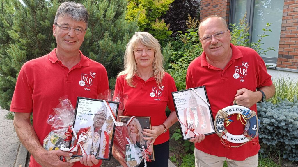 Zwei Männer und eine Frau in roten T-Shirts stehen vor einem Haus mit Büschen davor. Sie halten Bilder in der Hand.